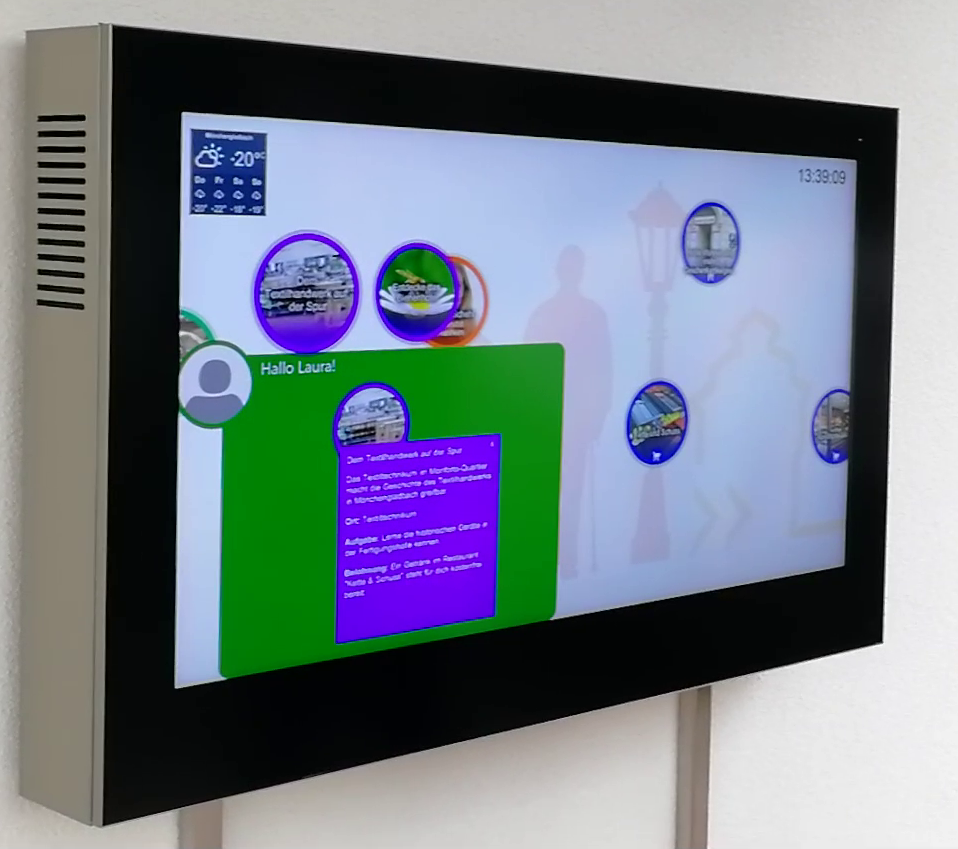 A large interactive touch screen visibly built for outdoor use (seemingly contained in a steel bezel) is mounted to a wall. The screen shows a user's personal pinboard in green in the bottom left corner. On the pinboard there is a text that greets the user by name and a summary of their currently selected activity. Outside of the personal pinboard area, other activities are floating around as bubbles. The top corners of the screen show a weather report and a digital clock.