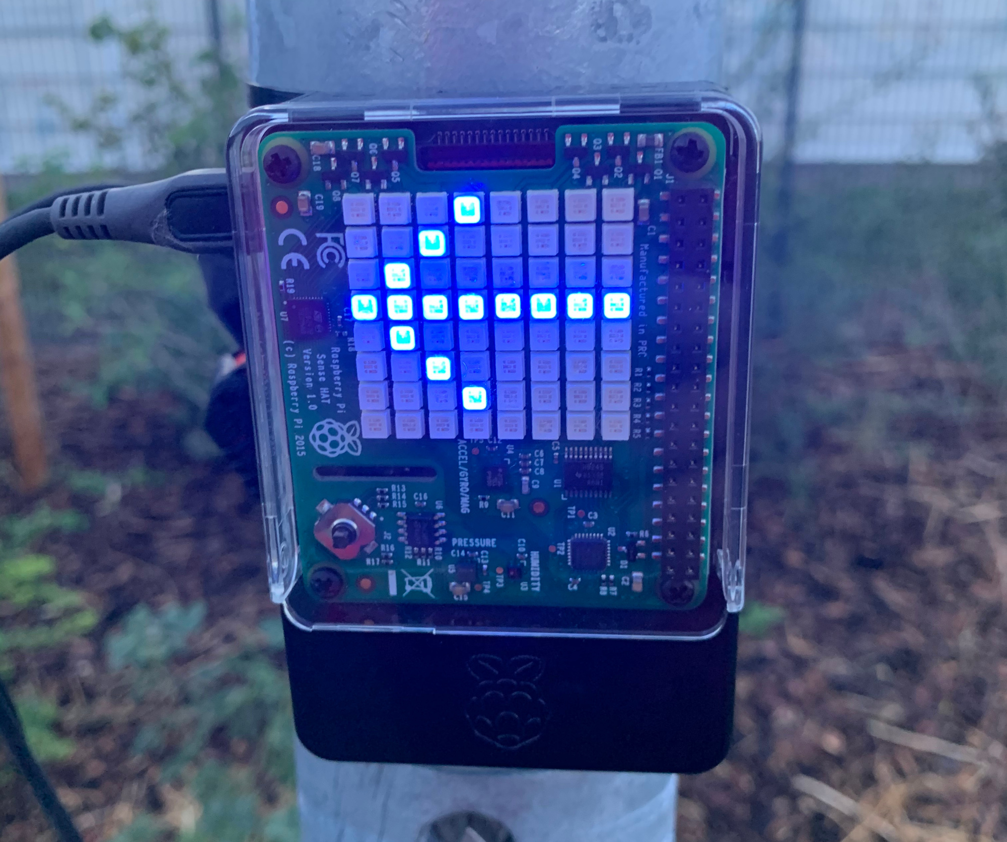 A close-up photo of a small computer with an LED matrix display which is currently showing a blue arrow pointing left. The computer is affixed to a steel pole. A flower bed is visible in the background.