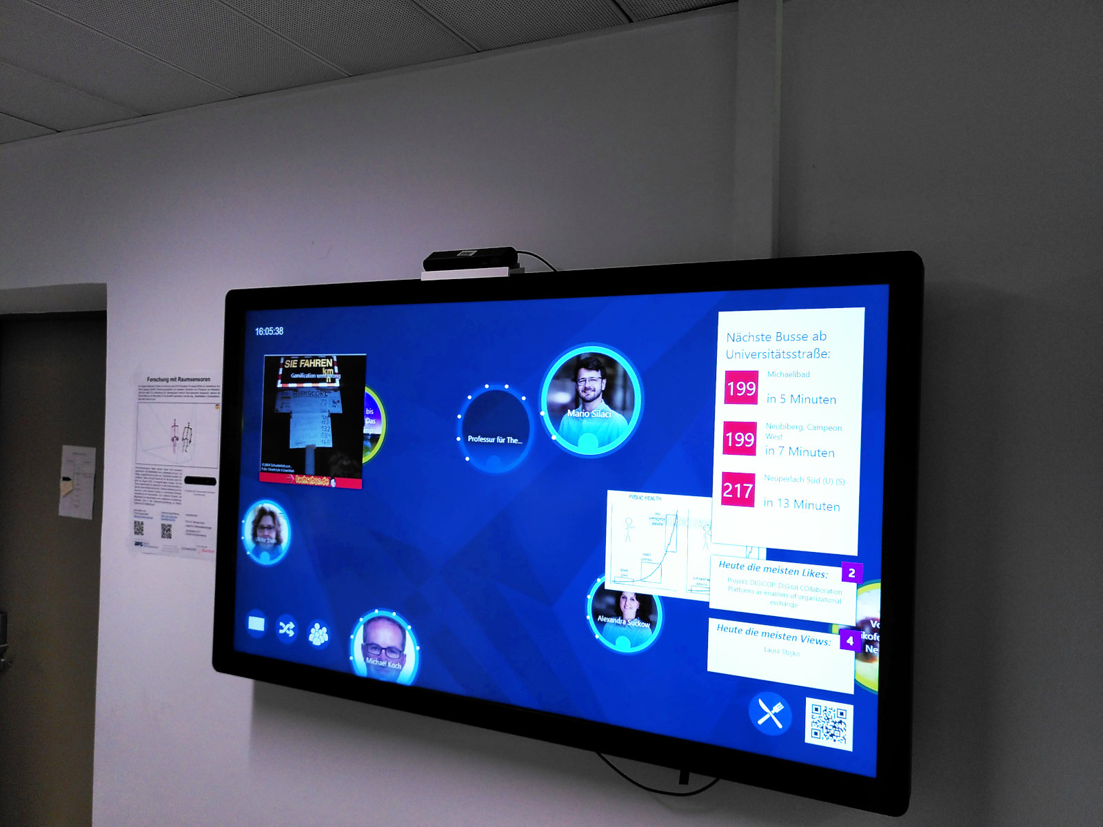A large touch screen mounted to a white wall in an indistinct office building, a door visible to its left