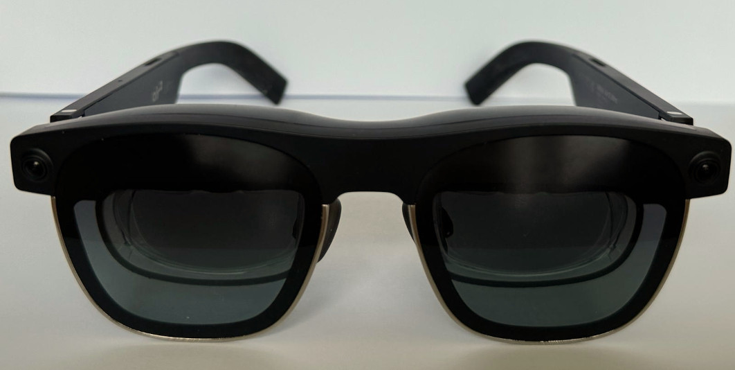 A paitr of smart glasses seen from the front. They look similar to normal black sunglasses, but have a thicker frame and what looks like two layers of glass.